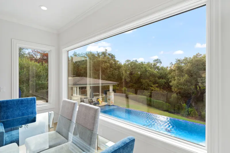 Wide white-framed picture window over an infinity-edge pool and oak trees